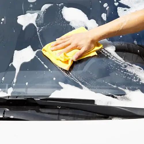 A hand using a yellow microfiber cloth to clean a car windshield with soap suds, showcasing Ultra Soft Microfiber Cleaning Cloths For Car Detailing Pack Of 24 Clean Without Scratch Lint Free Streak Free Absorbent Non Abrasive Reusable And Washable In Blue White Yellow By Amazon Basics.