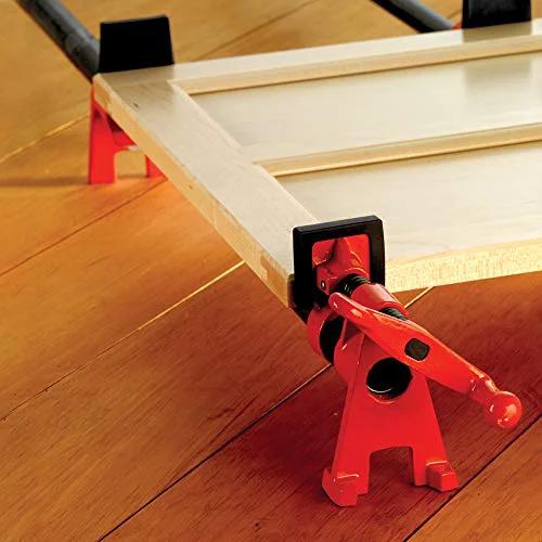 Red Versatile 3/4 Inch H Style Pipe Clamp in use for edge gluing wood panel, featuring stable dual-axis foot design on wooden floor.