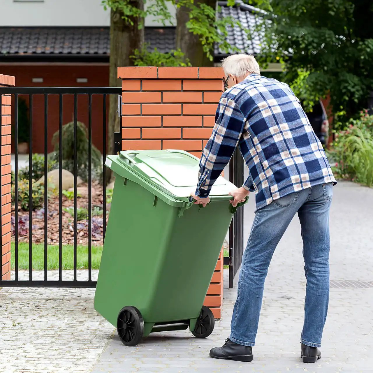 Person moving green garbage can with black wheels, demonstrating Garbage Can Wheels Replacement With Metal Hollow Shaft And Small Rubber Wheels 19.68 X 7.28 X 3.93 Inches Quiet Trash Can Pulley For Easy Installation And Efficient Waste Collection in a garden setting.