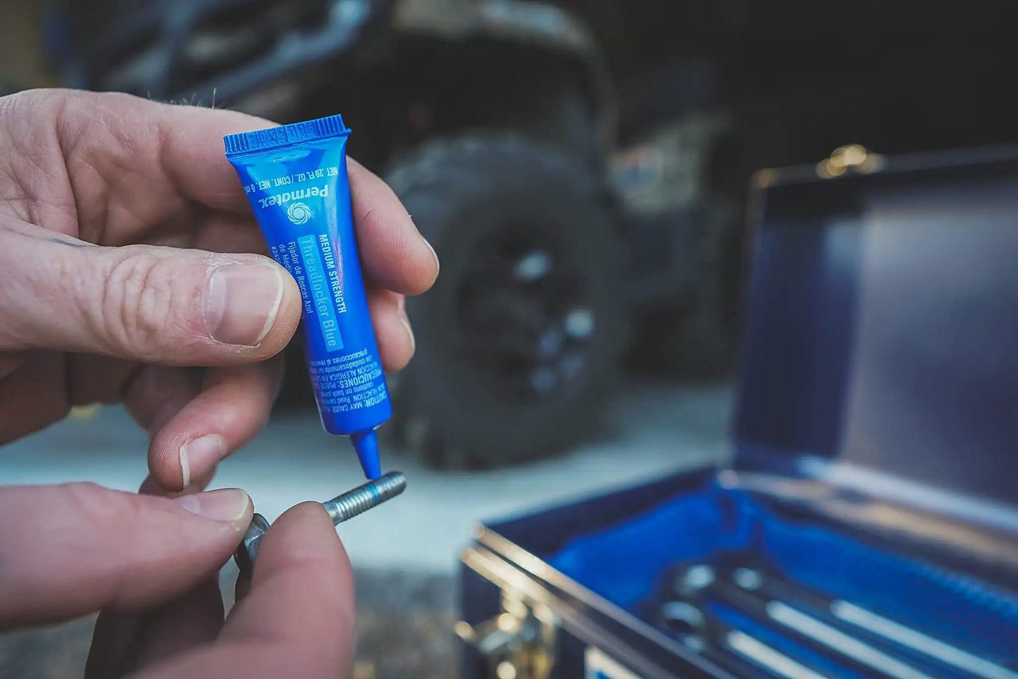 Person applying Blue Medium Strength Threadlocker That Eliminates Vibration No Drip Seals Leaks Prevents Rust And Disassembles Without Cracking Or Shrinking Permatex 27 070 6 Ml to a metal bolt with a toolbox in background.