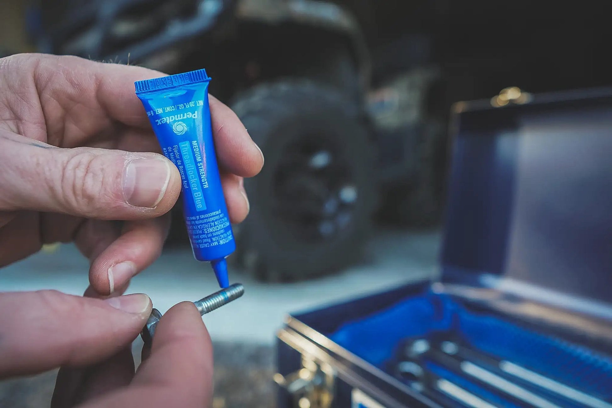 Person applying Blue Medium Strength Threadlocker That Eliminates Vibration No Drip Seals Leaks Prevents Rust And Disassembles Without Cracking Or Shrinking Permatex 27 070 6 Ml to a metal bolt with a toolbox in background.