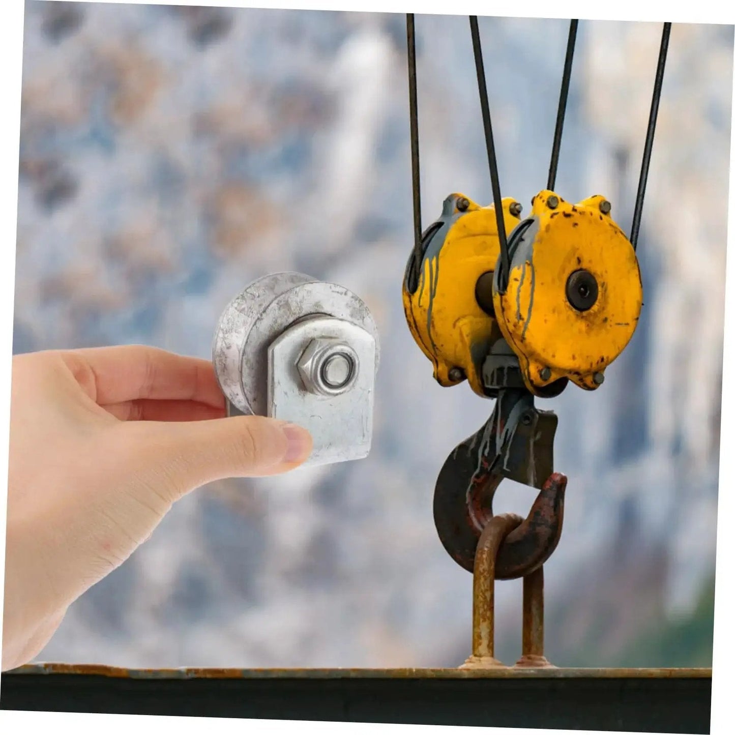 Hand holds a small metallic wheel next to a large industrial pulley system with yellow casings against a blurred background.