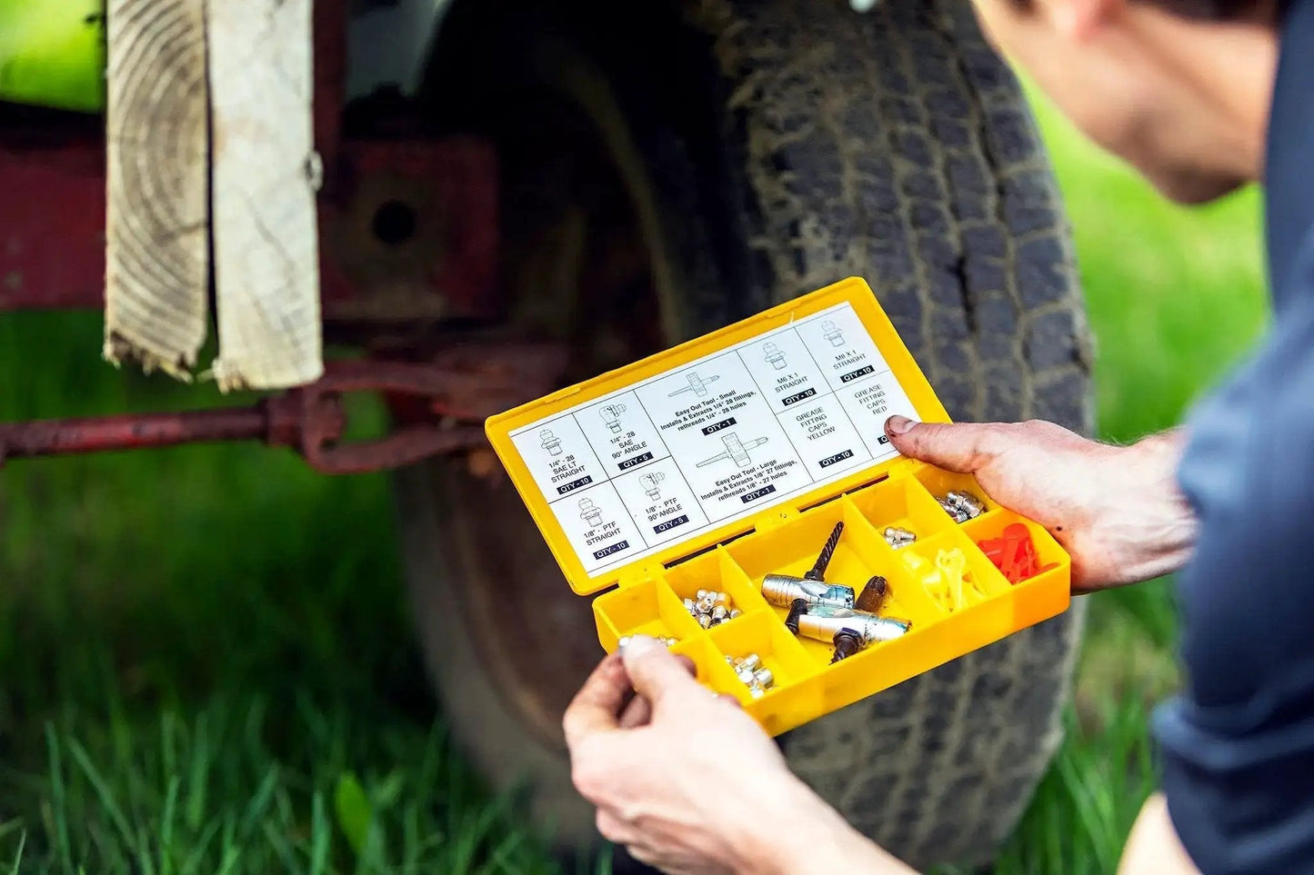 Person holding a Complete Grease Fitting Replacement Kit With SAE And Metric Zerks For Straight And Ninety Degree Fittings Ten Each Of One Eighth NPT One Quarter Twenty Eight SAE LT Six By One Metric Eight By One Metric Includes Two Multi Tools And Twenty Caps LockNLube near vehicle tire.