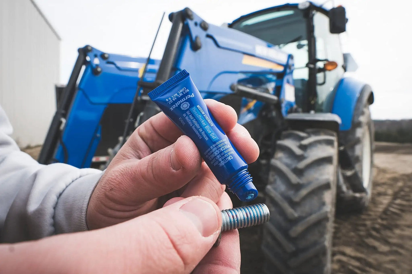Blue Medium Strength Threadlocker That Eliminates Vibration No Drip Seals Leaks Prevents Rust And Disassembles Without Cracking Or Shrinking Permatex 27 070 6 Ml held in hand with a metal bolt, tractor in background.