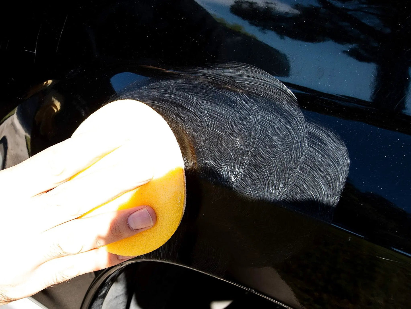 Hand applying Meguiars ScratchX 2.0 G10307C scratch remover for car paint, using a yellow applicator on a black car surface.