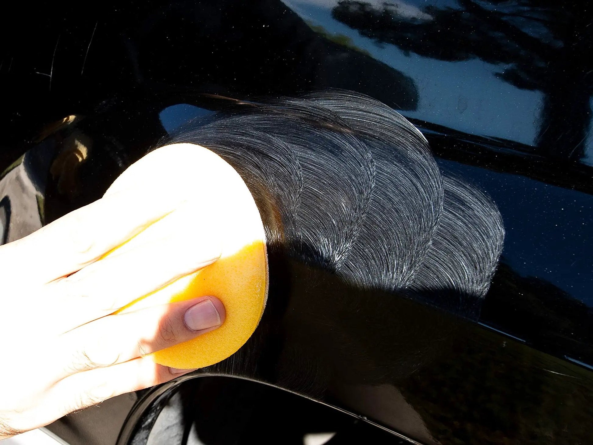 Hand applying Meguiars ScratchX 2.0 G10307C scratch remover for car paint, using a yellow applicator on a black car surface.