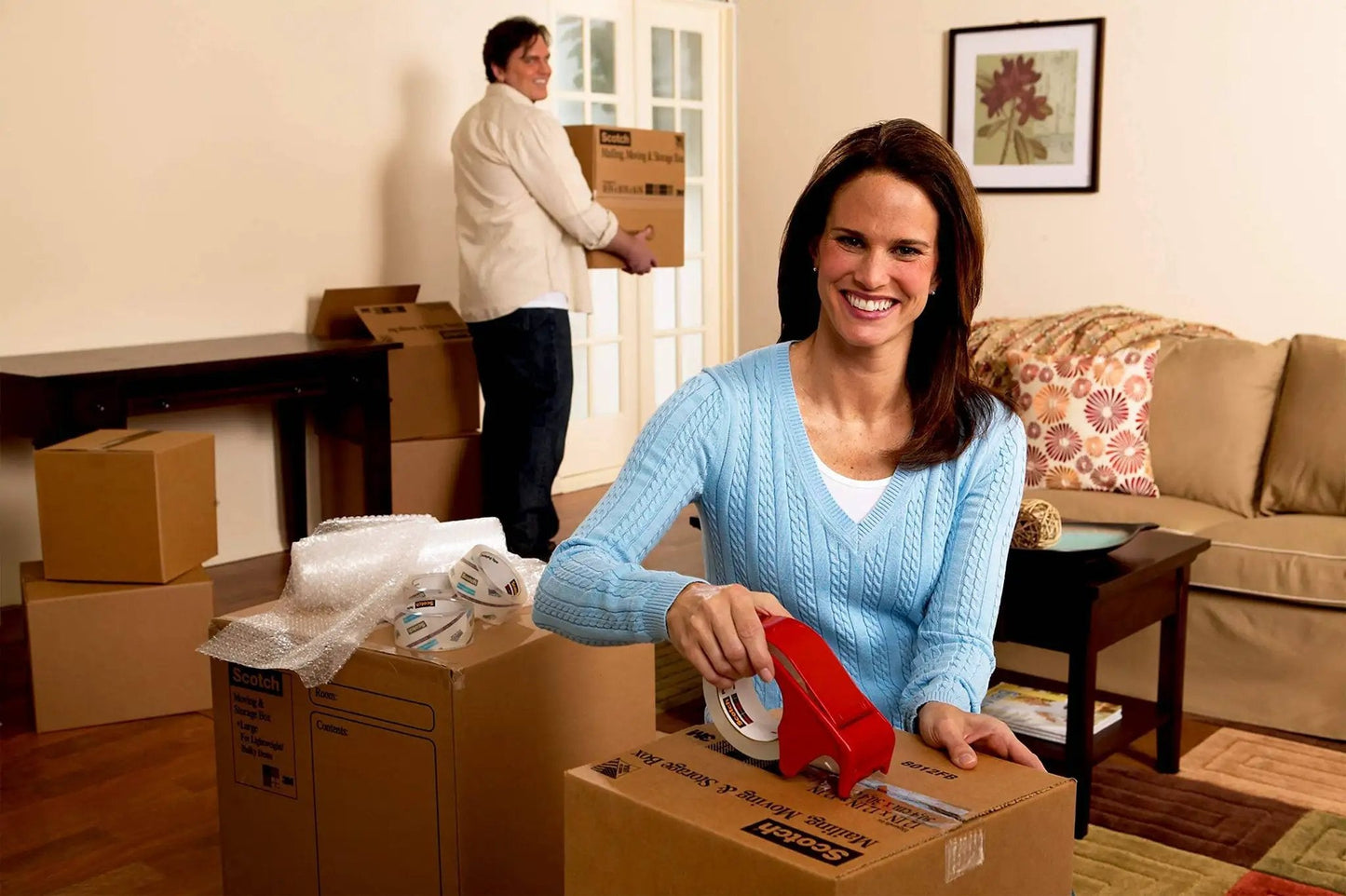Woman using portable heavy-duty packaging tape dispenser with hand brake for 3 in core rolls up to 2 in wide in a living room setting.