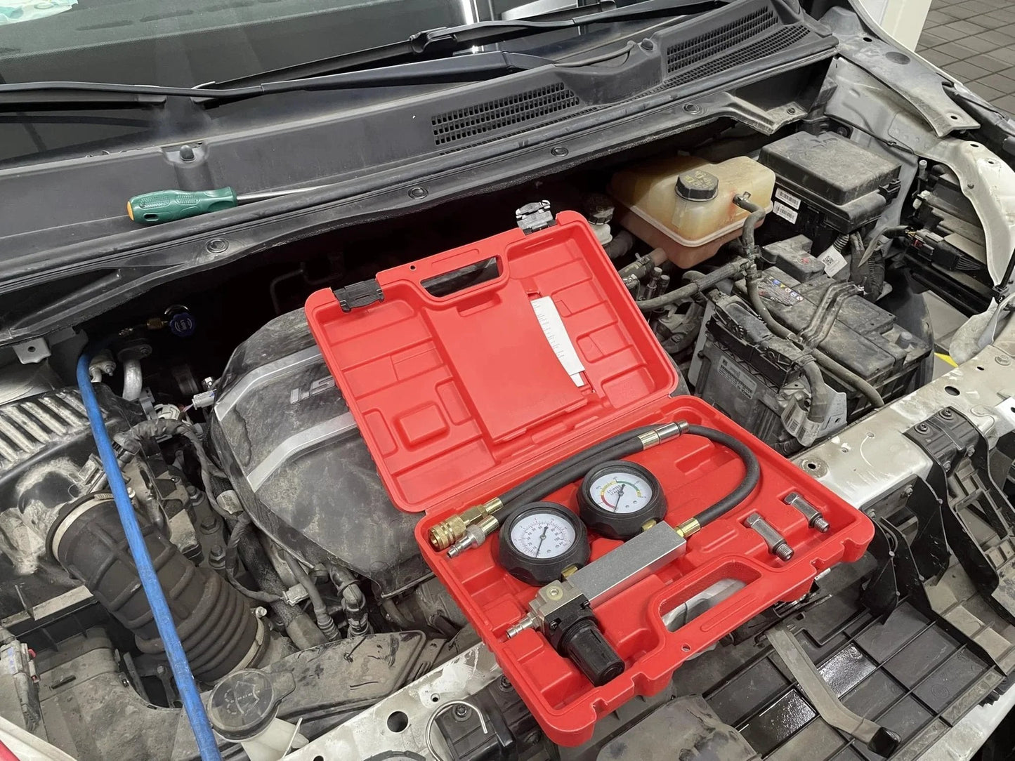 Cylinder Leak Detector Kit For Engine Compression Diagnosis And Cylinder Leakage Testing With 480mm Hose 12mm And 14mm Spark Plug Adapters 0 To 100 PSI Gauge Air Regulator Carrying Case HFS in open red case near car engine.