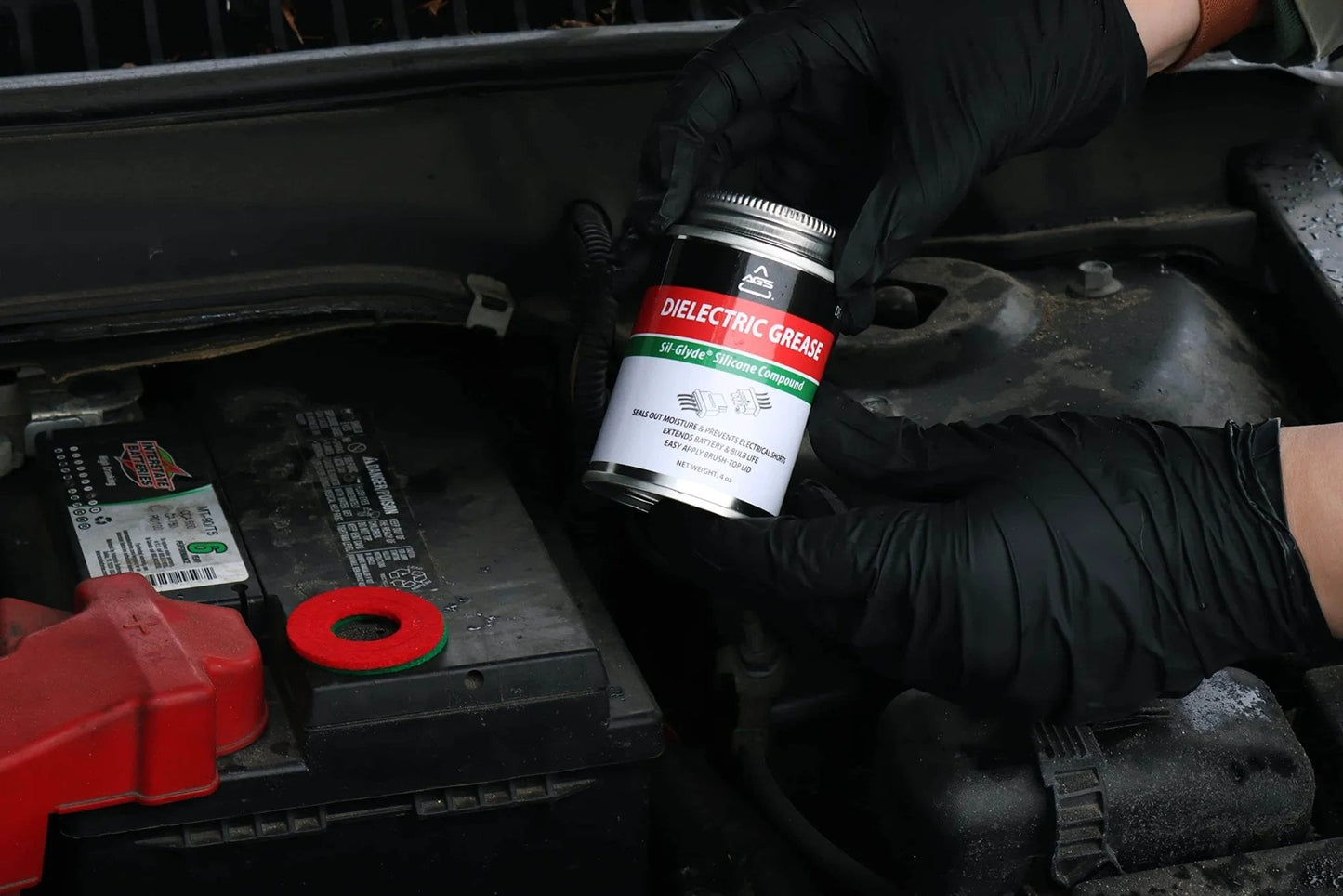 AGS Automotive Solutions Brush Top Can Dielectric Silicone Grease Compound for All Electrical Components and Connectors, 4oz being held near car battery