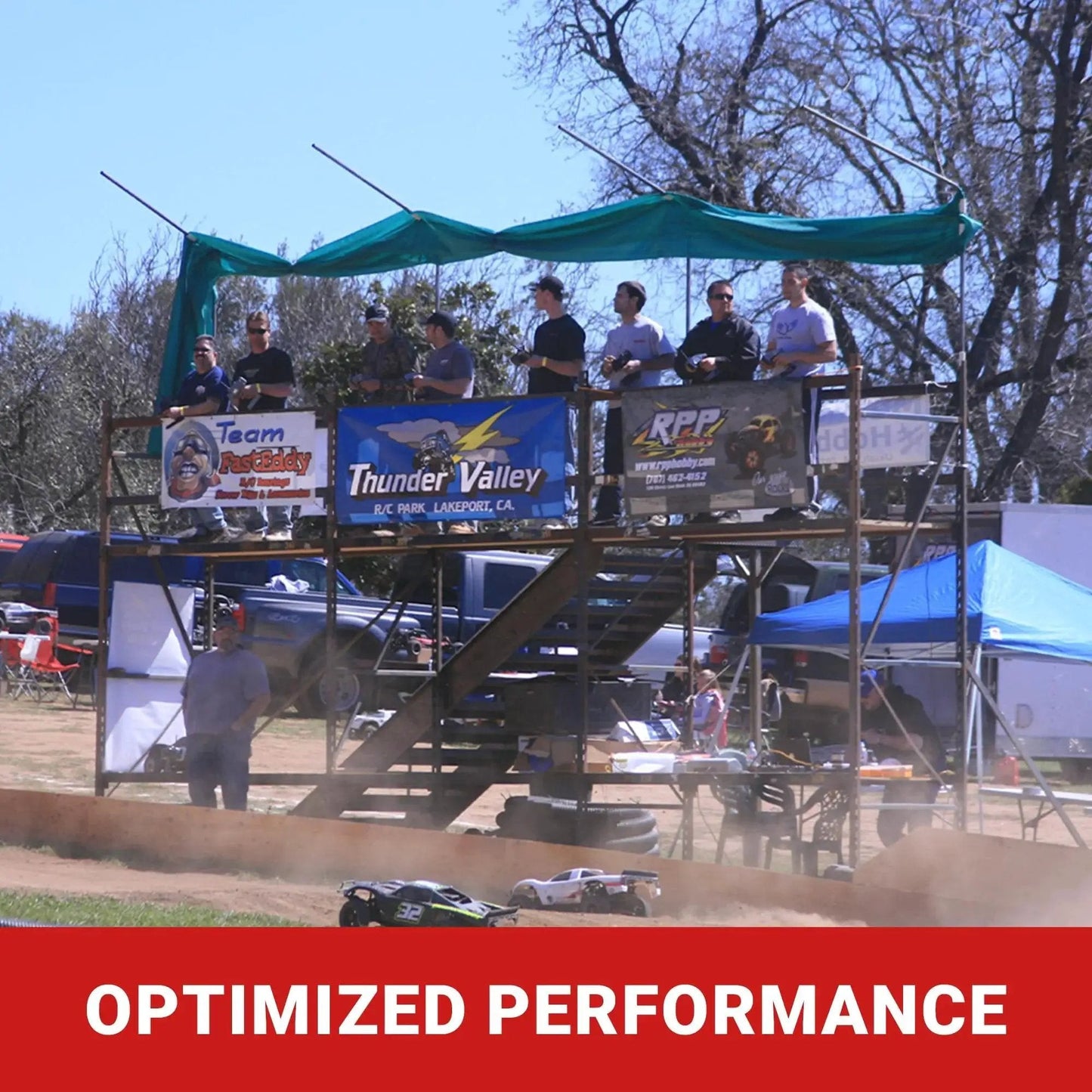 RC racers on platform at off-road event with banners and RC trucks in action below.